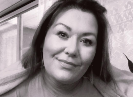 Black and white portrait of an adult smiling gently at the camera, sitting indoors with a window in the background.