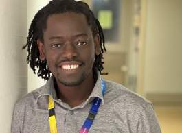 Man of color in a gray long sleeve shirt, leaning against the wall, smiling