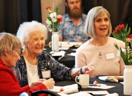 Three individuals at a social gathering seated around a table adorned with flowers, engaging in a cheerful conversation. One person is listening intently while another is speaking animatedly, with desserts on their plates.