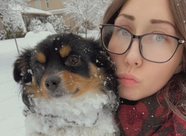 A young person with big brown glasses and brown hair with pursed lips holds a black, brown and white dog with a snowy scene in the background