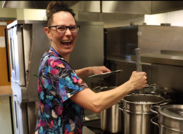 a woman smiles and laughs while mixing food in large pots at a kitchen in a hospital