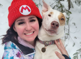 Woman with brown hair wearing red toque with letter “M” on it holds a white dog with a brown spot around its eye. There are snow and pine trees in the background. 