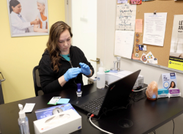 A person wearing blue medical gloves performs a test while sitting at a desk in front of a laptop in an office with yellow walls.