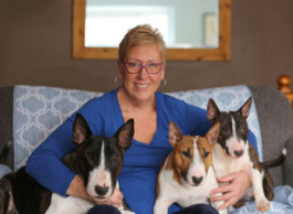 A smiling woman with short hair and glasses looks to the camera while sitting on a couch with her three dogs. 