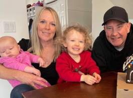 Smiling long-haired blonde woman holds baby in pink onesie, toddler in red top, seated at wood table beside smiling man wearing black ballcap and hoodie.