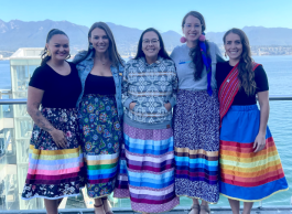 Five people standing arm in arm wearing brightly coloured ribbon skirts posing in front of ocean and mountains.