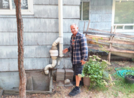 A person in a blue checked coat and black shorts points to white pipes outside a grey clapboard home