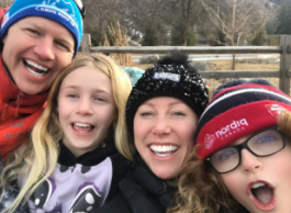 Four people smiling for a group selfie outdoors, with three wearing toques. Background includes a wooden fence and leafless trees.