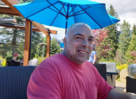 A smiling person sits in a chair on a public patio on a sunny day.