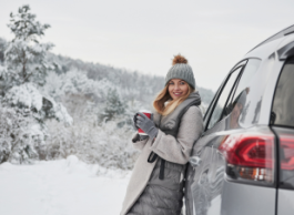 A young blonde woman holds a coffee and leans on her grey SUV while outside in winter near trees.