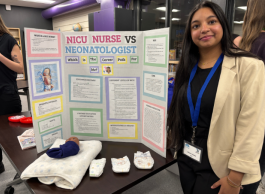Person standing next to a science fair poster titled "NICU Nurse vs Neonatologist," detailing career paths and roles, in a room with people in the background.