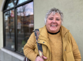 A smiling person with grey hair wearing a yellow winter jacket and sweater and carrying a purse