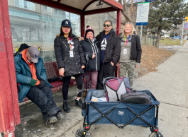 Five individuals stand and sit at a bus stop; one person is napping, covered in a blanket. A cart with a blue bag is beside them. A sign boasting "abc" and another sign with bus information are present in the background.