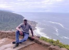 Man sitting on a rock wall, smiling with the ocean and coastline behind him.
