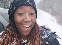 Smiling woman wearing dark jacket and toque hat covered in snowflakes.
