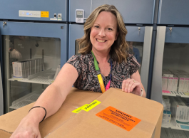 A smiling person with sandy blonde hair and short-sleeved blouse and lanyard opens a cardboard box in front of a series of blue fridges.
