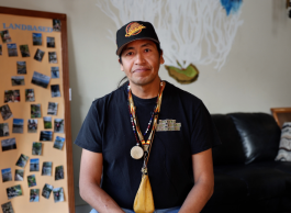 A person in a ballcap that says Canucks and a black tshirt with necklaces around their neck sits on a black leather chair next to a cupboard adorned with photographs.