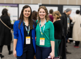 Two people stand side by side at a trade show or conference. They are both wearing lanyards and one person is wearing a green sweater, the other a blue jacket.