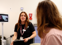 A person with long brown hair and pink shirt with their back to the camera watches a nurse wearing a black tshirt and lanyard. There's a screen in the background with a person wearing headphones.