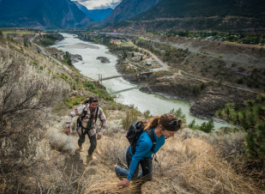 Two people hike up a bushy hill overlooking a large body of water with houses on either side connected by a bridge.