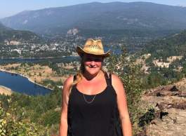 Smiling, long-haired blonde woman wearing cowboy hat, black sleeveless top and necklace stands on top of hill overlooking lake and mountain range.