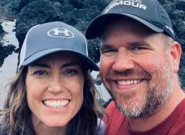 Man in grey cap and red shirt with woman in blue cap and long dark hair standing together smiling in front of a waterfall.