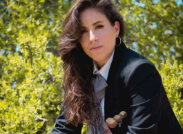 A woman with long dark curled hair and hooped earrings is sitting on a rock with greenery in the background. She is dressed as El Charro Negro, wearing a traditional Mexican black charro suit and a wide-brimmed hat, a red tie around her waist with gold tassels up her legs and on her breast pocket.