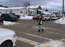 A person in a high-vis safety vest, jeans, botts and pink toque holds up a stop sign at a busy intersection on a snowy day.