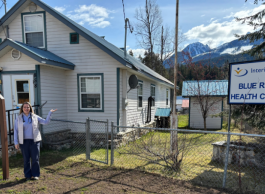 A person in a white vest and blue shirt stands in front of an old white home with a green roof. A sign to the right says Blue River Health Centre. Snowcapped mountains and blue sky are in the background.