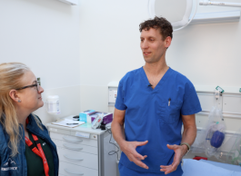 A man in blue medical clothing speaks to a woman with glasses and long blonde hair tied back in a a pony tail. They are in an exam room in a hospital.