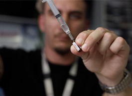 A man with facial hair and short blonde hair wearing a black t-shirt and lanyard holds up a syringe and a vial.
