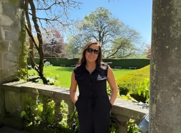 Woman with dark brown hair wearing sunglasses and a black outfit stands between stone columns, in front of a colourful garden with plants and trees. 
