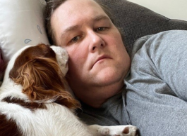 A person wearing a grey top lies on a pillow on a couch next to a brown and white dog.