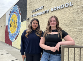 Two teenaged girls both with long blond-brown hair and dark tops stand next to a railing in front of a building with a mural that says Lillooet Secondary School.