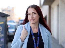 A woman with long red-brown hair wearing a light blue business jacket, dark shirt and lanyard holds a purse over her shoulder as she walks down a city street.