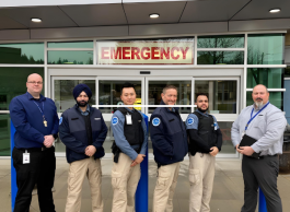 A group of men of different ages and ethnicities wearing security uniforms stand with their arms crossed in front of doors with a sign that says EMERGENCY above them.
