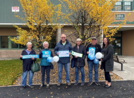 A group of six people of varying ages wearing warm clothing stand with certificates and balloons in hand. They are standing in front of trees changing colours with a building in the background that says Community Health.