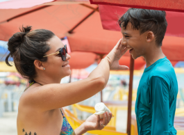 A woman with her hair in a bun and wearing sunglasses and bathing suit applies sunscreen to a smiling boy wearing a teal coloured shirt under a red umbrella.