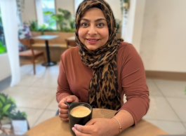 a woman wearing a headscarf and smiling is sitting at a table drinking a cup of coffee