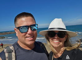 Two people wearing sunglasses smile while talking a selfie on the beach near the water. One person has a backpack strap over their shoulder and the other is wearing a white sun hat.