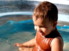 A very young child with short brown hair wearing an orange swimsuit splashes in a wading pool on a sunny day.