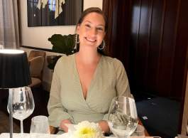 Smiling woman with brown hair wearing hoop earrings and gold V-neck top with white dots sits at table, with beige chairs and brown wood panel door in background.