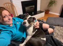 A selfie of a brown-haired person wearing a blue sweater relaxing indoors with a multi-coloured dog beside them.