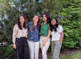 A group of four people in business casual clothing posing in front of a stand of pine and deciduous trees.