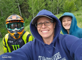 Three people stand shoulder to shoulder to take a selfie style picture in a very green forest setting. One with a biking helmet, and the other two with their sweater hoods up.
