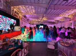 A group of people dancing on a multi-coloured floor next to a lit up stage in a ballroom.