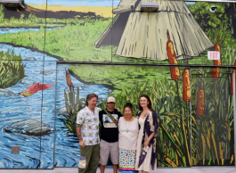 A group of four people laughing stand arm in arm in front of a large mural depicting a river, salmon, bullrushes and lodge on grass.