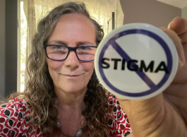 A person with long curly grey hair and glasses holds up a button that has a slash through the word stigma.