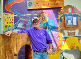 A person with a grey beard wearing a ball cap and purple shirt poses casually in a colourful playroom with a picture of a young person in a frame in the background.