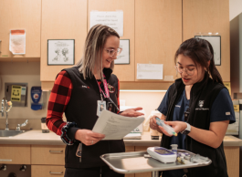 Two health-care professionals chat with one another as one examines instruments on a tray. They're standing in an examination room with cabinets and a sink with eye wash station.